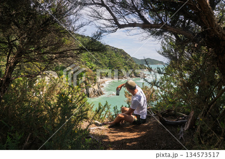 Hiker sitting in forest taking phone photo of coast, Abel Tasman, New Zealand 134573517