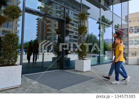 Couple walks towards a modern building with large glass windows on a sunny day in an urban setting Couple walks towards a modern building with large glass windows on a sunny day in an urban setting 134575385