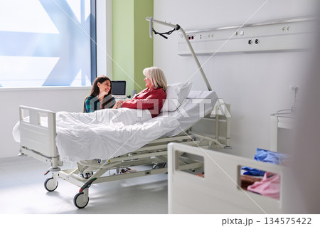Woman visits patient in hospital room during daytime, sharing a moment of care and support in a bright and clean environment 134575422