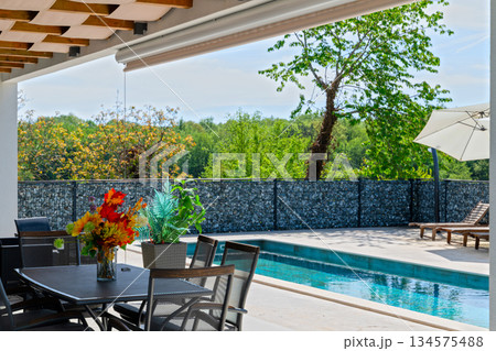 Covered terrace of a modern modular house with dining table, overlooking a swimming pool and gabion fence. Contemporary prefabricated home architecture with outdoor living space 134575488