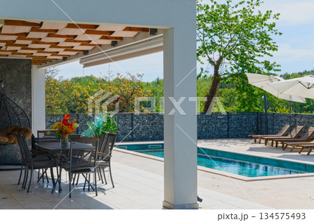 Covered terrace of a modern modular house with dining table, overlooking a swimming pool and gabion fence. Contemporary prefabricated home architecture with outdoor living space 134575493