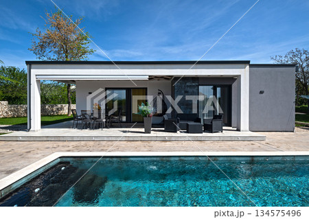 Modern modular house backyard featuring a rectangular swimming pool, large patio with wooden sun loungers, and umbrellas against a blue sky. Contemporary prefabricated home design Modern modular house backyard featuring a rectangular swimming pool, large patio with wooden sun loungers, and umbrellas against a blue sky. Contemporary prefabricated home design 134575496