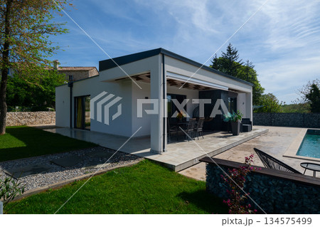 Covered terrace of a modern modular house with dining table, overlooking a swimming pool and gabion fence. Contemporary prefabricated home architecture with outdoor living space 134575499