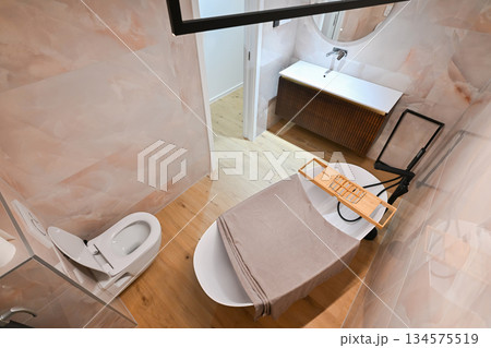 Luxurious modern bathroom featuring a sleek freestanding bathtub with a caddy, elegant vanity with sink and mirror, and a contemporary toilet. Soft tiled walls and wooden flooring create a spa-like 134575519