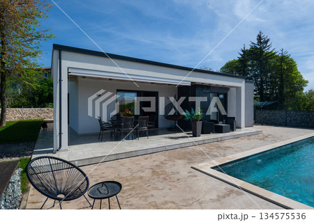 Modern modular house backyard featuring a rectangular swimming pool, large patio with wooden sun loungers, and umbrellas against a blue sky. Contemporary prefabricated home design Modern modular house backyard featuring a rectangular swimming pool, large patio with wooden sun loungers, and umbrellas against a blue sky. Contemporary prefabricated home design 134575536