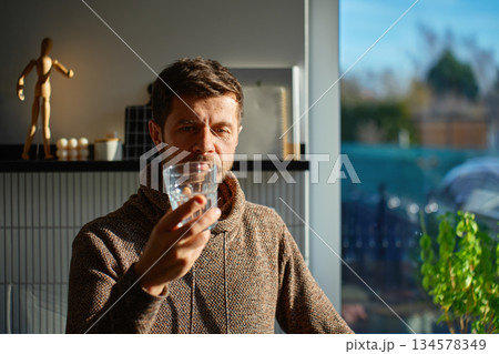 Man drinking water from glass in kitchen interior with daylight Man drinking water from glass in kitchen interior with daylight 134578349