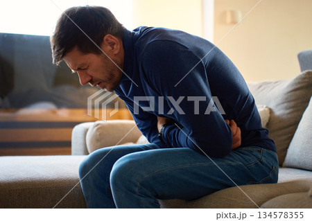 Man clutching his stomach, suffering from abdominal pain at home Man clutching his stomach, suffering from abdominal pain at home 134578355