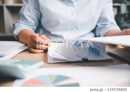 Office worker analyzing business reports and financial charts, reviewing data and writing notes to support planning, strategy and professional decision-making in a modern workplace environment. 134578809