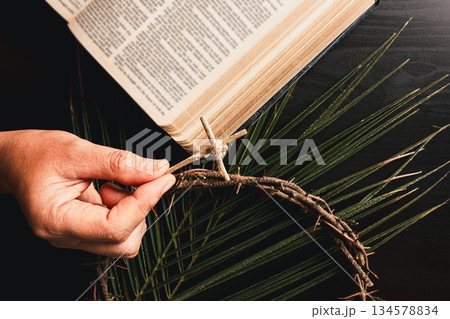 Hand attaching small palm leaf cross to crown of thorns above open Bible with palm leaves, symbolizing Christian faith, Palm Sunday ritual, sacrifice and Easter reflection for worship, prayer concepts Hand attaching small palm leaf cross to crown of thorns above open Bible with palm leaves, symbolizing Christian faith, Palm Sunday ritual, sacrifice and Easter reflection for worship, prayer concepts 134578834