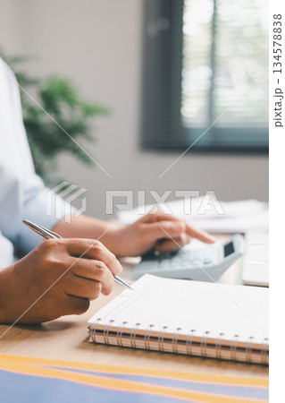 Close-up of office worker writing financial records in notebook while holding calculator, illustrating accounting tasks, expense tracking, budgeting and professional financial management in workplace 134578838
