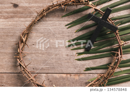 Black wooden cross with crown of thorns, palm leaves on rustic wooden table, symbolizing Christian faith, sacrifice, Palm Sunday, Easter reflection, religious background for worship. 134578848