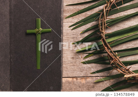 Palm leaves cross with crown of thorns, palm leaves beside Bible on rustic wooden table, symbolizing Christian faith, sacrifice, Palm Sunday, Easter reflection, religious background for worship. 134578849
