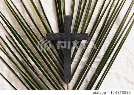 Wooden cross on green palm leaves with crown of thorns over white fabric background, symbolizing Christian faith, Palm Sunday, sacrifice, Easter reflection, minimal religious concept with copy space. 134578850