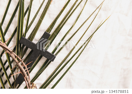 Wooden cross on green palm leaves with crown of thorns over white fabric background, symbolizing Christian faith, Palm Sunday, sacrifice, Easter reflection, minimal religious concept with copy space. 134578851