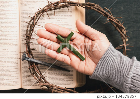 Hand holding palm leaf cross above open Bible, crown of thorns, iron nail, symbolizing Christian faith, Palm Sunday, sacrifice, Easter reflection, religious background for worship, prayer concepts. Hand holding palm leaf cross above open Bible, crown of thorns, iron nail, symbolizing Christian faith, Palm Sunday, sacrifice, Easter reflection, religious background for worship, prayer concepts. 134578857