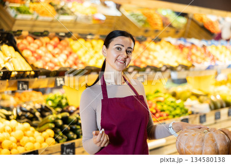 Portrait of positive female grocery supermarket employee near vegetables, fruits and pumpkins 134580138