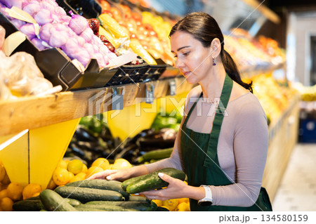 Female salesperson carefully places ripe cucumbers on counter of a grocery supermarket Female salesperson carefully places ripe cucumbers on counter of a grocery supermarket 134580159