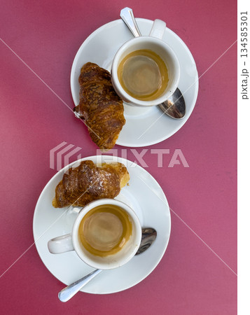 Two cups of espresso with croissants on white plates on a pink table setting Two cups of espresso with croissants on white plates on a pink table setting 134585301