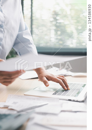 Office worker standing at desk while typing on laptop and reviewing multiple documents, illustrating business tasks, paperwork management, productivity and professional workflow in modern workplace. Office worker standing at desk while typing on laptop and reviewing multiple documents, illustrating business tasks, paperwork management, productivity and professional workflow in modern workplace. 134586930