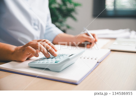 Close-up of office worker calculating financial figures with calculator while writing notes in notebook, illustrating budgeting, expense management, accounting tasks, professional financial workflow. 134586955