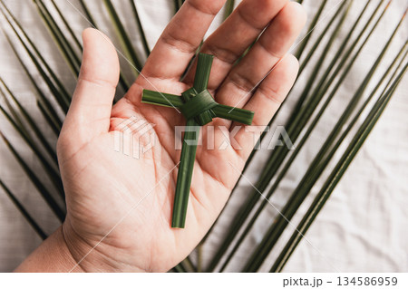 Hand holding palm leaf cross over white fabric and green palm leaves, symbolizing Christian faith, Palm Sunday, worship and spiritual devotion, minimal religious concept with natural light background. 134586959