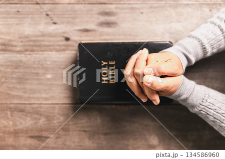 Hands in prayer above Holy Bible on wooden table, symbolizing Christian faith, worship, devotion, trust in God, spiritual religious background with copy space for church, prayer, meditation concepts. 134586960