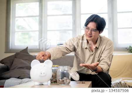 Asian office worker man putting coin into piggy bank aside glass jars sitting on sofa in living room Asian office worker man putting coin into piggy bank aside glass jars sitting on sofa in living room 134587506