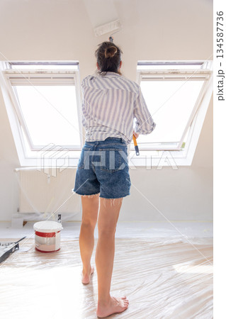 Back view young adult beautiful single happy woman painting wall paint roller brush white color living room moving new home flat . Female painter decorating interior renovation residential apartment 134587736