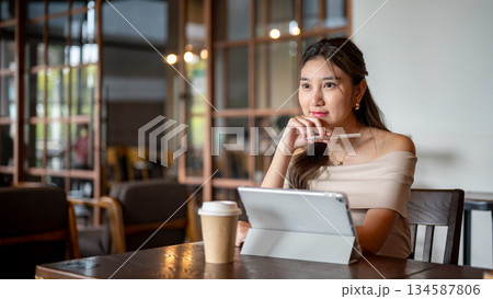 Asian woman looking ahead while sitting with tablet and coffee aside at table in cafe or coffee shop 134587806