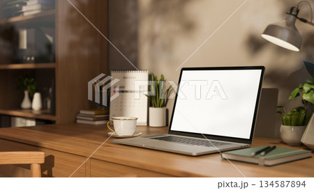 White screen laptop and coffee with lamp and calendar on wooden table aside bookshelf in study room. 134587894