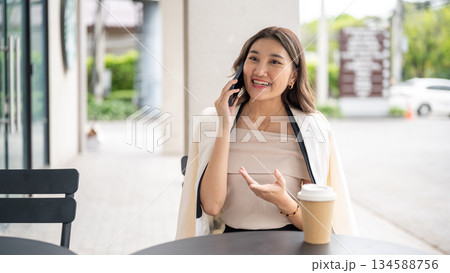 Asian woman smiling with explaining hand as talking on the phone with coffee cup on cafe black table Asian woman smiling with explaining hand as talking on the phone with coffee cup on cafe black table 134588756