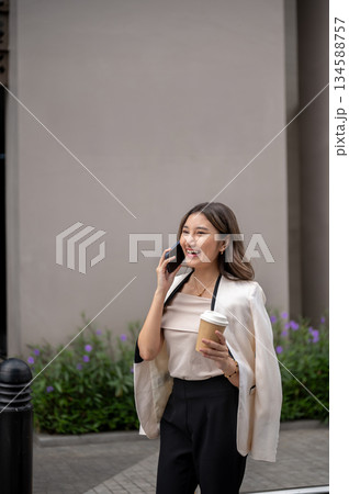 Long hair asian woman talking on phone while walking on sidewalk around building with coffee in hand Long hair asian woman talking on phone while walking on sidewalk around building with coffee in hand 134588757