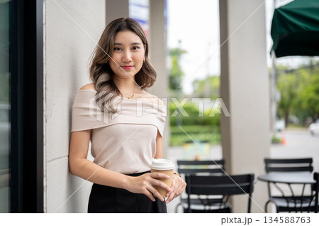 Long hair asian woman standing near a building's grey wall and holding a coffee cup with both hands. 134588763