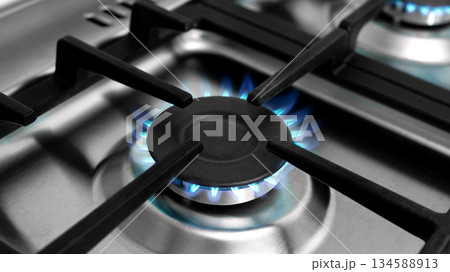 Close-up of a gas burner with blue flame on a stainless steel stove. Energy usage concept. Close-up of a gas burner with blue flame on a stainless steel stove. Energy usage concept. 134588913
