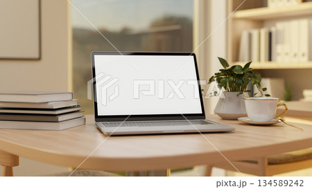 Blank screen laptop and coffee or tea cup on wooden table in sunlit study room with bookshelf. 134589242