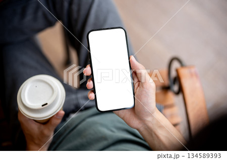 Close up of a hand holding blank screen smartphone reading news with a coffee cup on wooden bench. 134589393