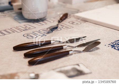 Set of three artist palette knives with wooden handles placed on a textured surface, surrounded by art supplies, showcasing tools for painting and creativity in a vibrant workspace 134589760