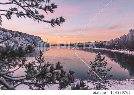 Serene winter landscape featuring a tranquil river reflecting pastel sunset hues, surrounded by snow-covered trees and a peaceful atmosphere, ideal for nature lovers and relaxation themes 134589765