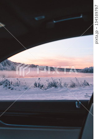 Scenic winter landscape viewed through car window, showcasing snow-covered terrain, frosty trees, and a serene river, with soft pastel colors of dawn illuminating the sky 134589767