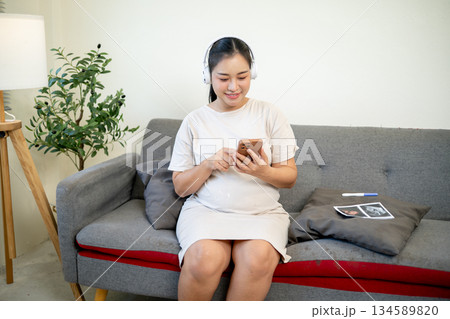Pregnant asian woman wearing headphones listening to classical music while sitting on sofa in house. Pregnant asian woman wearing headphones listening to classical music while sitting on sofa in house. 134589820