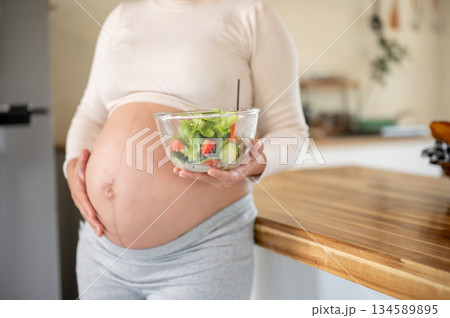 Close up of pregnant woman holding a glass bowl of vegetable salad and touching her belly in kitchen Close up of pregnant woman holding a glass bowl of vegetable salad and touching her belly in kitchen 134589895
