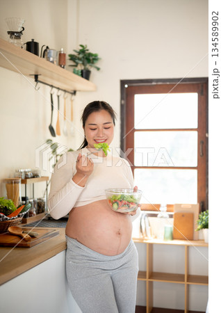 Pregnant asian woman holding a glass bowl enjoy eating a vegetables salad while standing in kitchen. 134589902