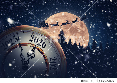 Magical holiday time transition to 2026. A snowy, New Year's Eve clock is set against a massive full moon with Santa Claus and his reindeer flying across the dark, festive sky. 134592805