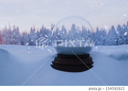 Empty Snow Globe at Sunset. Crystal ball on a snowy hill, with a backdrop of a frosted pine forest silhouette against a soft pink and orange sky. Ideal for winter and New Year concepts. 134592821
