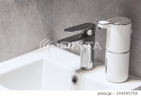 Close-up of a chrome faucet and white touchless liquid soap dispenser in a minimalist bathroom interior. The concept of hygiene, cleanliness, modern smart home technology, and hand washing. Close-up of a chrome faucet and white touchless liquid soap dispenser in a minimalist bathroom interior. The concept of hygiene, cleanliness, modern smart home technology, and hand washing. 134595758