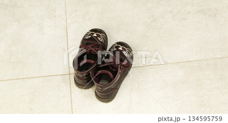 A pair of burgundy leather children's boots with a cute cat design, standing on light-colored tiles. Top view, concept of childhood, coming home, fashion for kids, and family comfort in the interior. A pair of burgundy leather children's boots with a cute cat design, standing on light-colored tiles. Top view, concept of childhood, coming home, fashion for kids, and family comfort in the interior. 134595759
