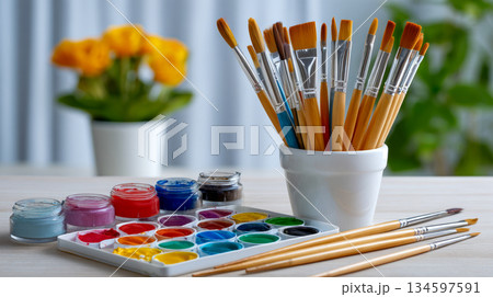 A vibrant array of art supplies is on a table, A palette with various watercolors and a set of paintbrushes in a holder A vibrant array of art supplies is on a table, A palette with various watercolors and a set of paintbrushes in a holder 134597591