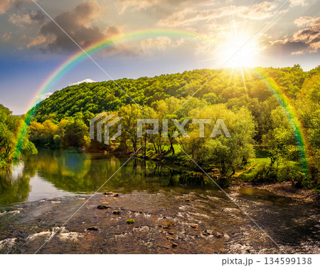 River flows among green forest near mountain at sunset. picturesque nature of a serene spring landscape in evening light. deciduous trees reflection. timeless nostalgia book cover with rainbow River flows among green forest near mountain at sunset. picturesque nature of a serene spring landscape in evening light. deciduous trees reflection. timeless nostalgia book cover with rainbow 134599138