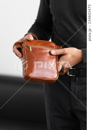 Male model in a charcoal knit crewneck and tailored trousers wearing a cognac leather vertical messenger bag against a white studio background. Male model in a charcoal knit crewneck and tailored trousers wearing a cognac leather vertical messenger bag against a white studio background. 134603473