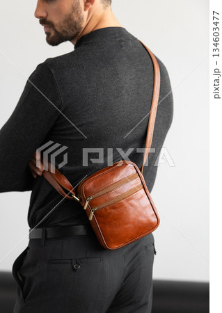 A stylish man in a dark grey crew neck sweater poses with a cognac leather crossbody bag against a minimalist white studio background. A stylish man in a dark grey crew neck sweater poses with a cognac leather crossbody bag against a minimalist white studio background. 134603477